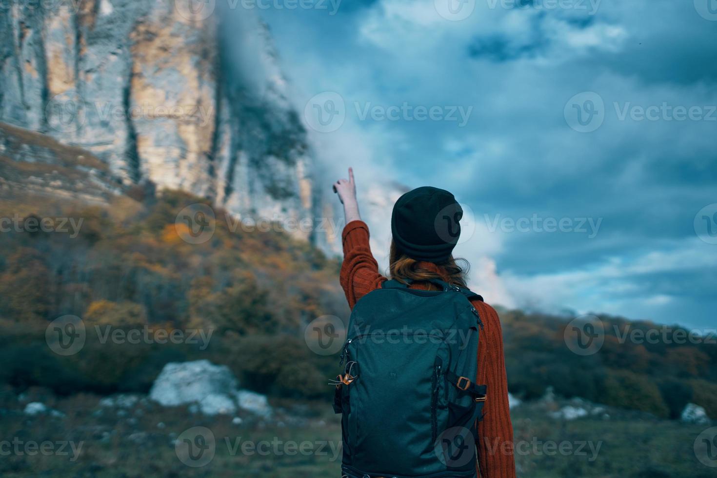 a traveler with a backpack and in a red sweater shows the mountains with her hand and the blue sky clouds photo