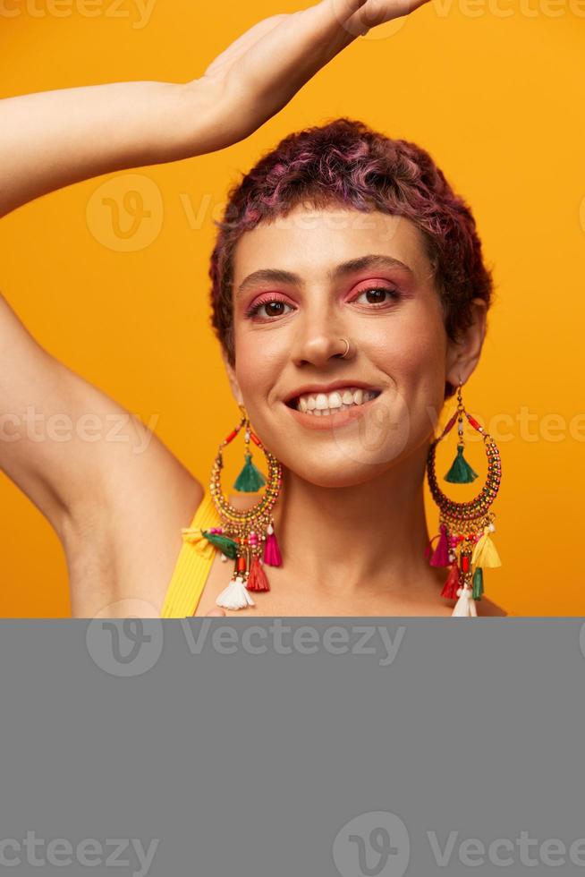 Portrait of a young woman with a short haircut and colored hair smiling and showing her tongue at the camera on an orange background with earrings accessories in the studio photo