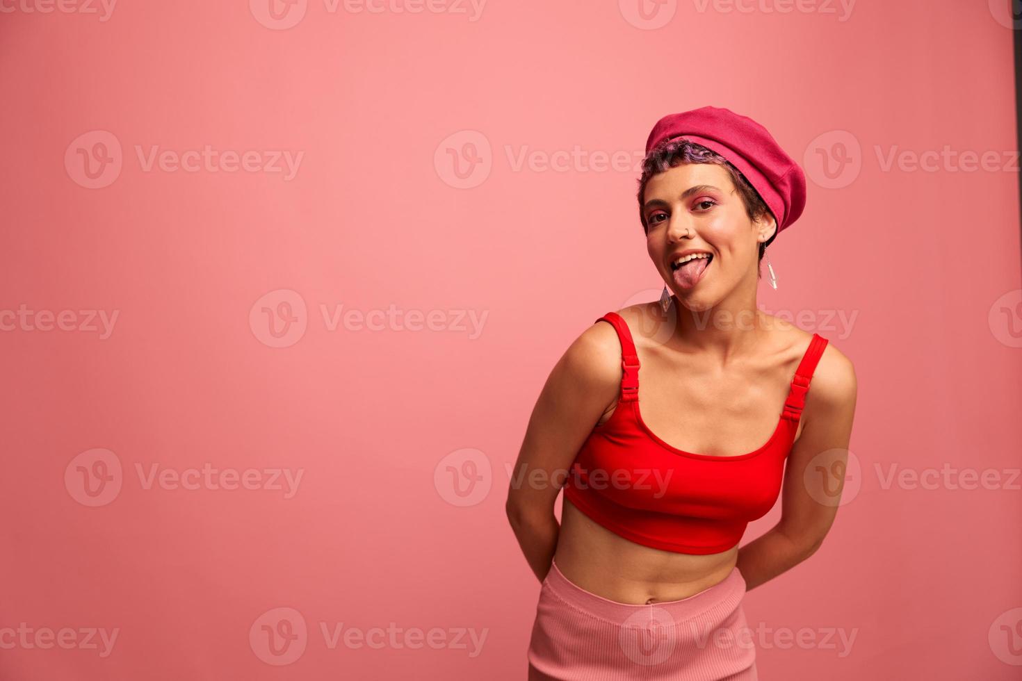 Young athletic woman with a short haircut and purple hair in a red top and a pink hat with an athletic figure smiles and grimaces looking at the camera on a pink background photo