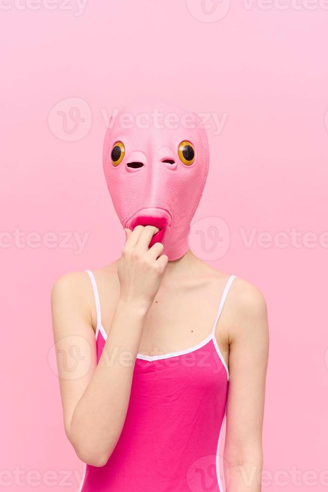 A funny woman wearing a pink fish mask on her head put her fingers in her mouth with pink clothes on a pink background. The concept of modern art photography photo