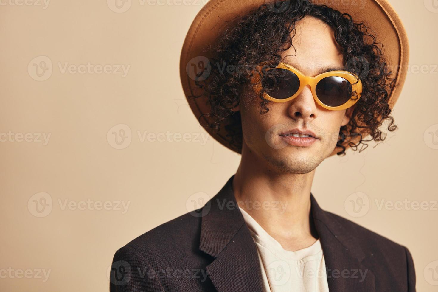 Cute awesome tanned curly man in classic hat brown jacket trendy sunglasses posing isolated on beige pastel background. Fashion New Collection offer. Retro style portrait concept. Free place for ad photo