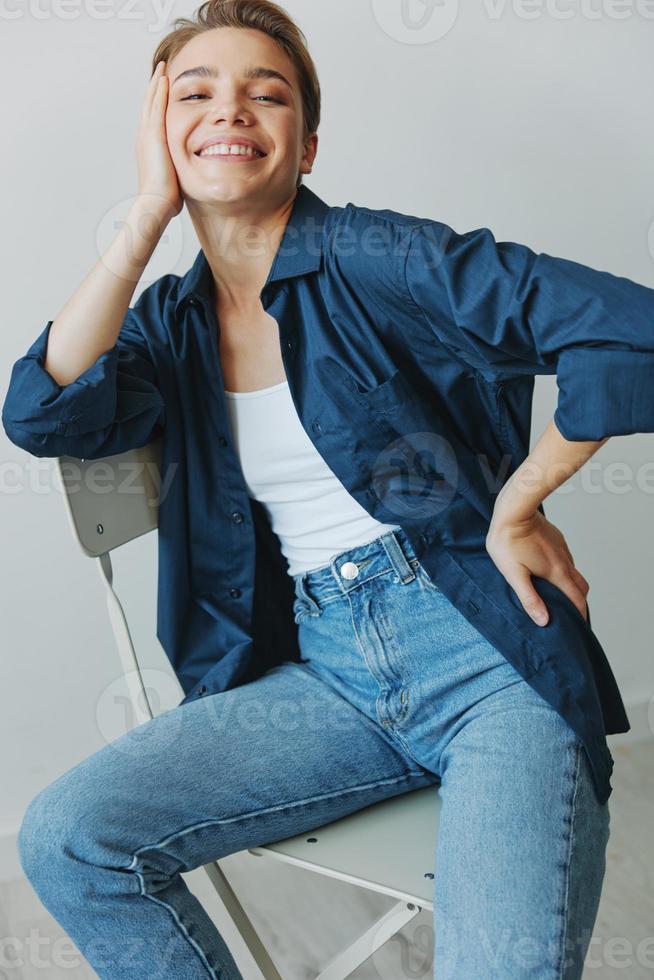 un joven mujer sentado en un silla a hogar sonriente con dientes con un corto Corte de pelo en pantalones y un mezclilla camisa en un blanco antecedentes. niña natural poses con No filtros foto