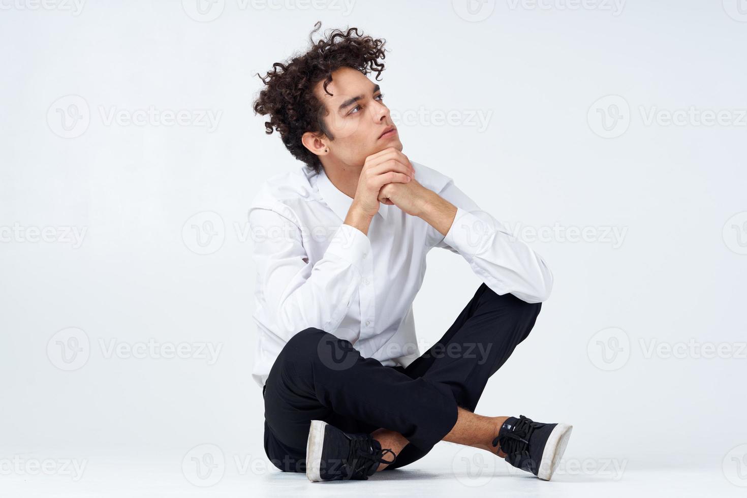 chico con Rizado pelo en un blanco camisa, zapatillas y pantalones se sienta en el piso foto