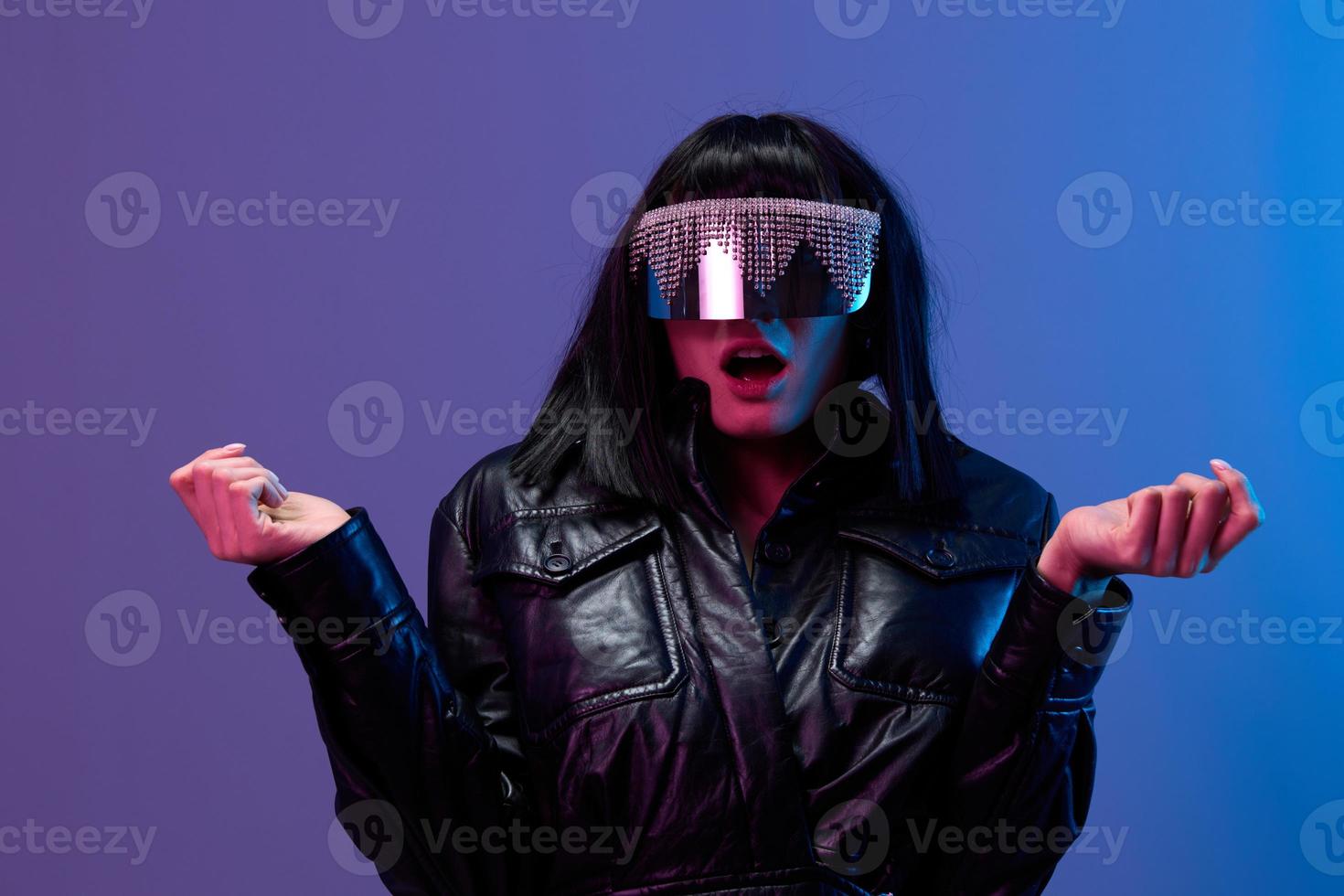 confuso conmocionado bonito moreno dama en cuero chaqueta de moda de espejo Gafas de sol abierto boca untado manos posando aislado en azul Violeta color ligero antecedentes. neón fiesta cyberpunk concepto. Copiar espacio foto