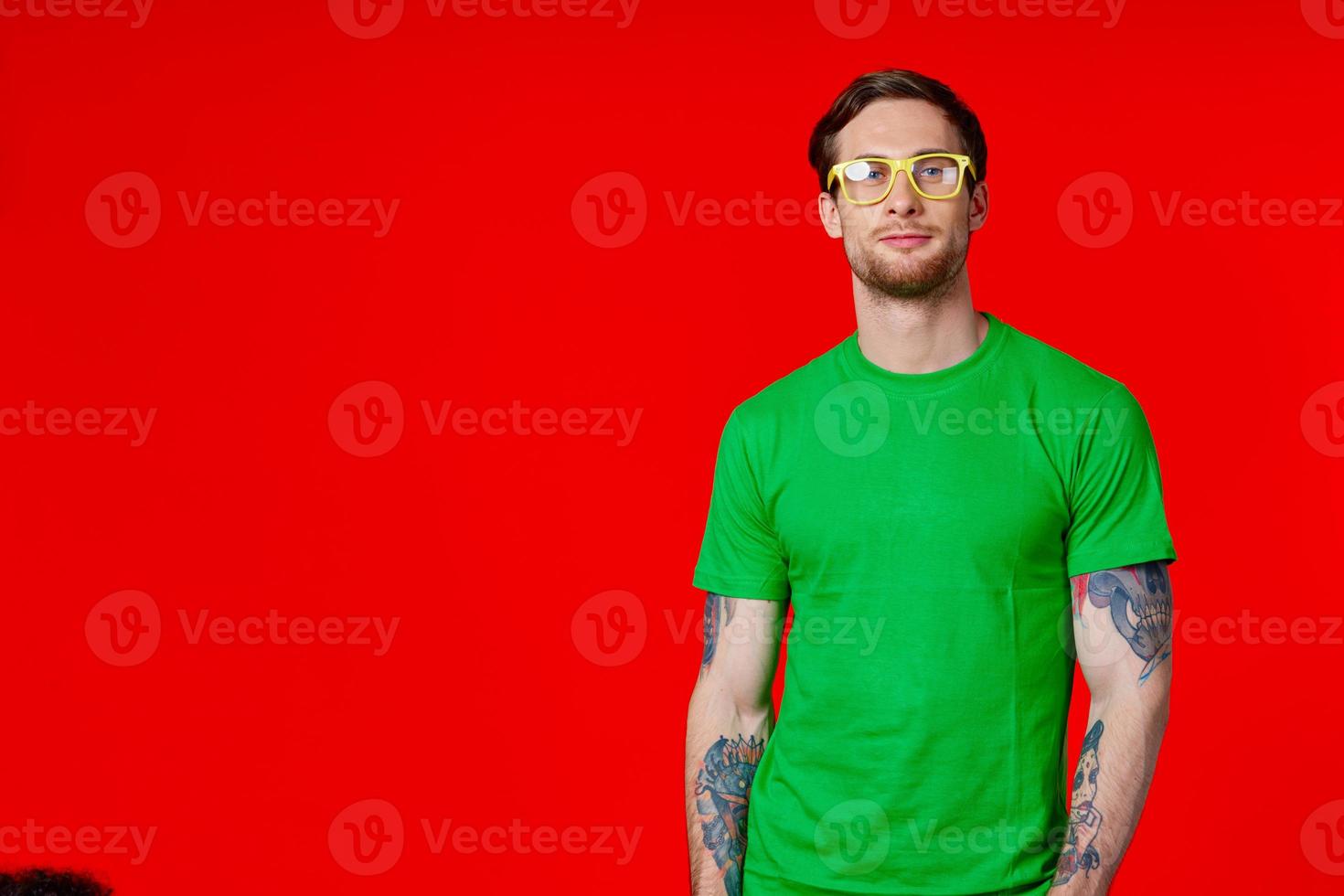 a man in a green t-shirt with tattoos in his hands wearing glasses on a red background photo