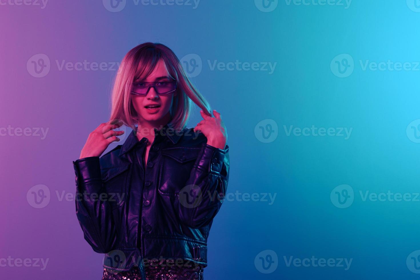 Flirting sexy adorable beautiful blonde woman in leather jacket sparkly dress trendy sunglasses posing isolated in blue pink color light studio background. Neon party Cyberpunk concept. Copy space photo