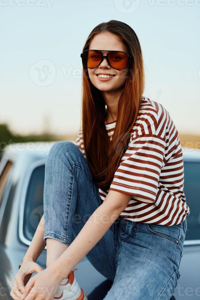A fashion woman in stylish sunglasses, a striped t-shirt and jeans sits on the trunk of a car and looks at the beautiful nature of autumn. Travel lifestyle photo