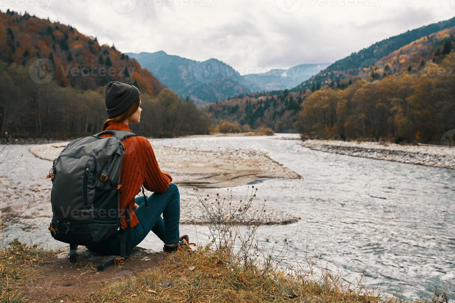 woman with a backpack in a jacket and a hat on the river bank in the mountains side view photo