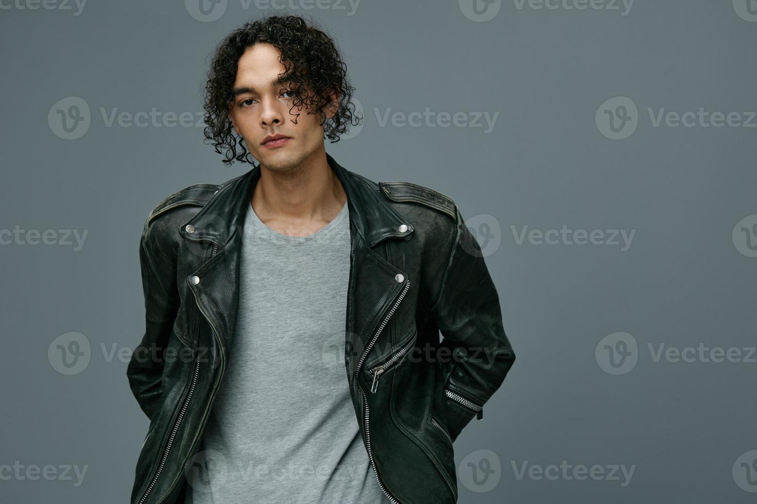 Self-confident stylish tanned curly man leather jacket looks at camera posing isolated on over gray studio background. Cool fashion offer. Huge Seasonal Sale New Collection concept. Copy space for ad photo