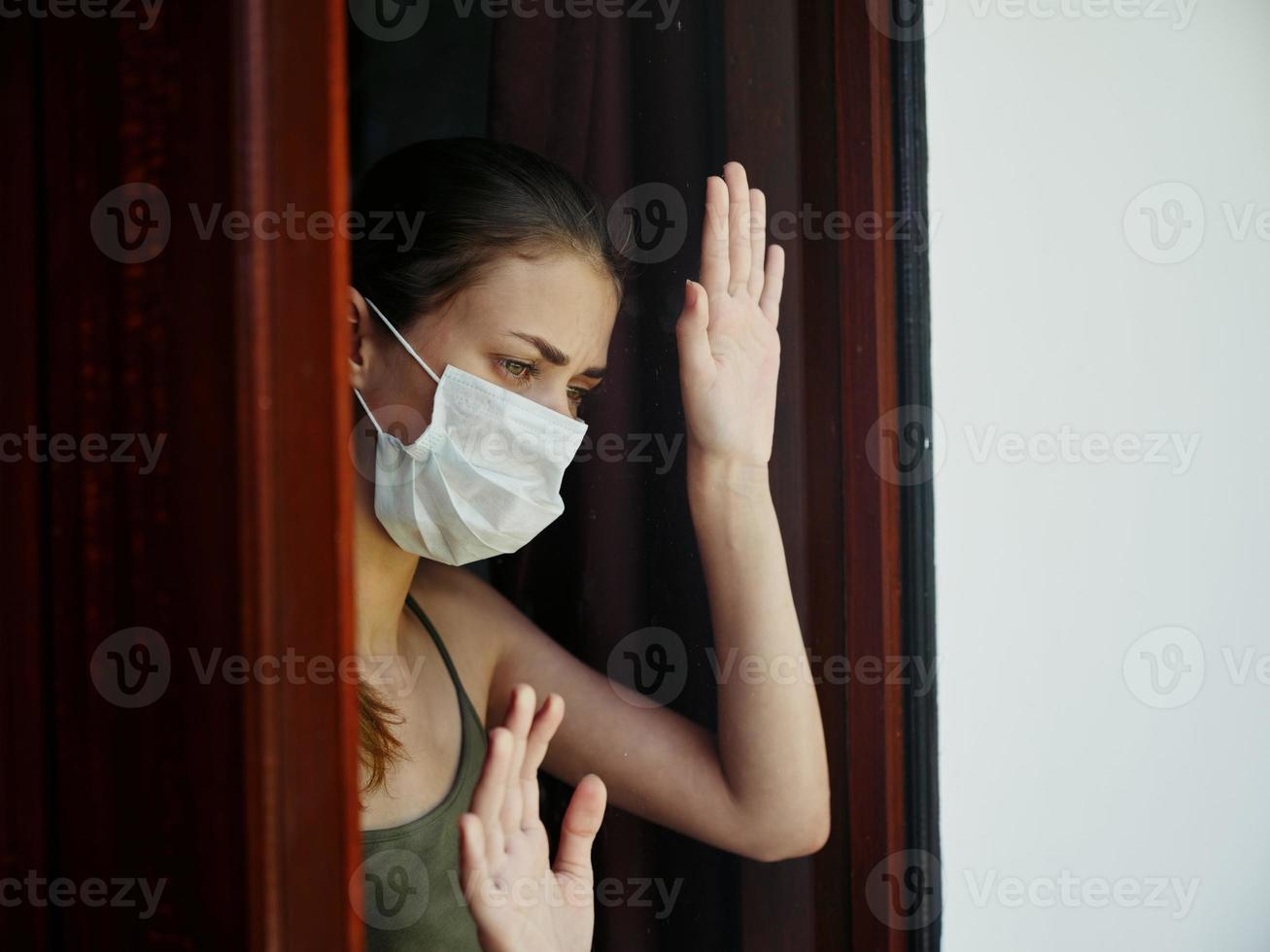 woman with sad facial expression in medical mask looking out the lockdown window photo