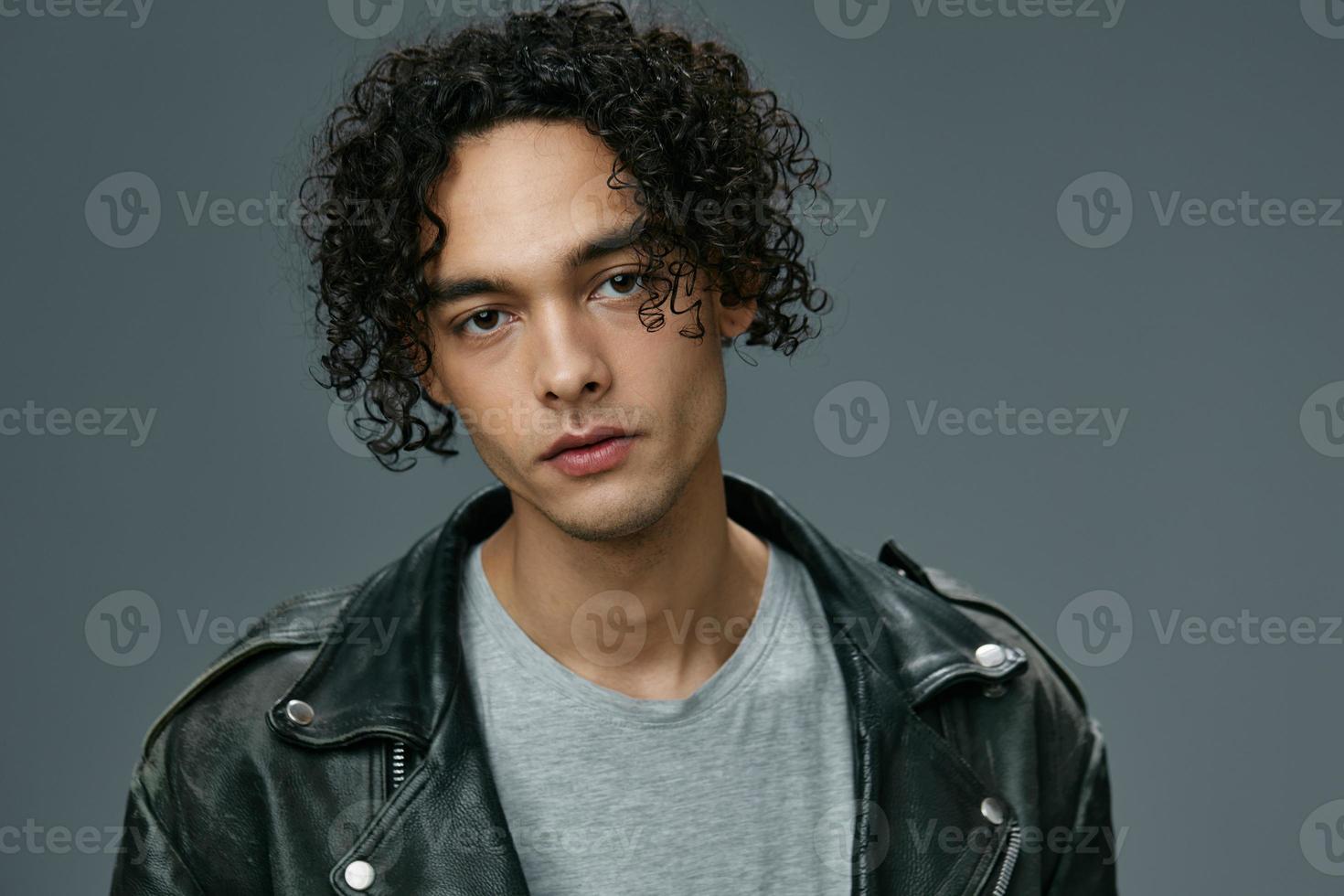Close up headshot portrait of stylish tanned curly man leather jacket posing isolated on over gray studio background. Cool fashion offer. Huge Seasonal Sale New Collection concept. Copy space for ad photo