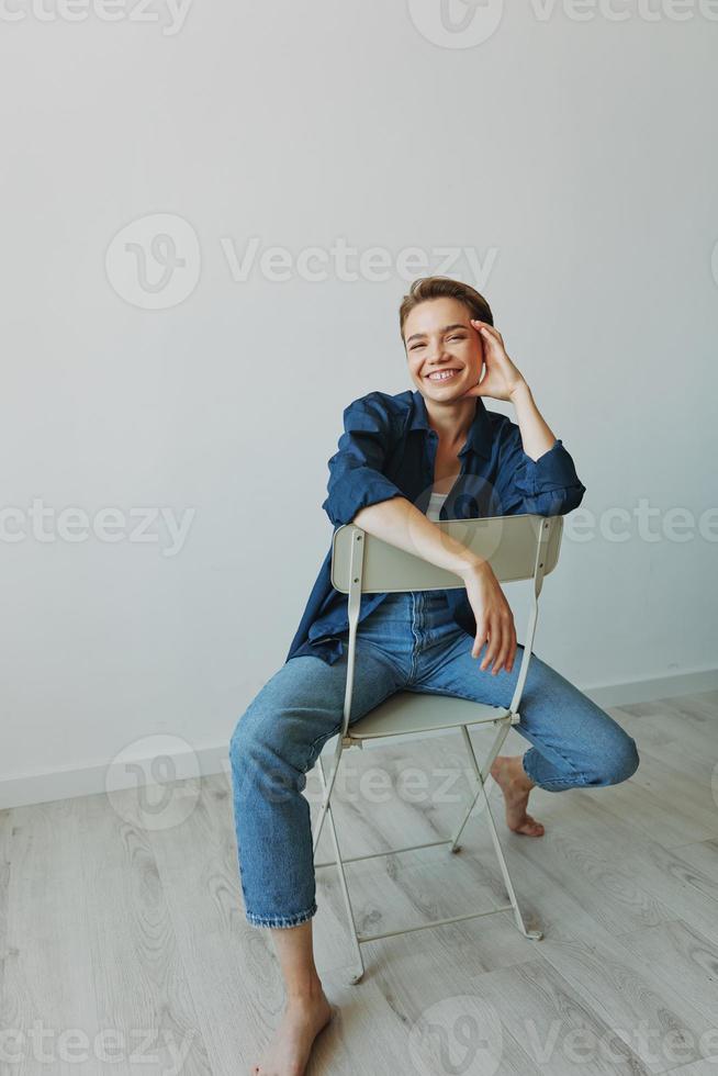 un joven mujer sentado en un silla a hogar sonriente con dientes con un corto Corte de pelo en pantalones y un mezclilla camisa en un blanco antecedentes. niña natural poses con No filtros foto