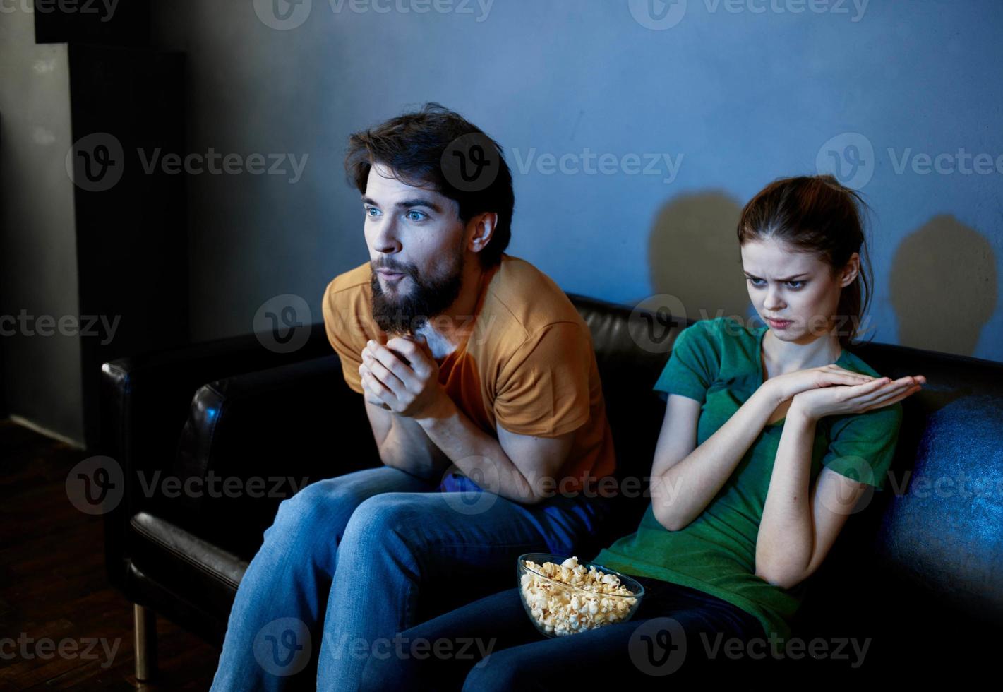 A woman and a man with popcorn on a leather sofa indoors watching TV in the evening photo