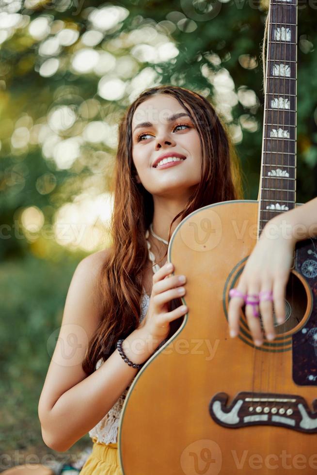 A young hippie woman with a guitar in her hands smiles sweetly into the camera on a trip to nature lifestyle in harmony photo