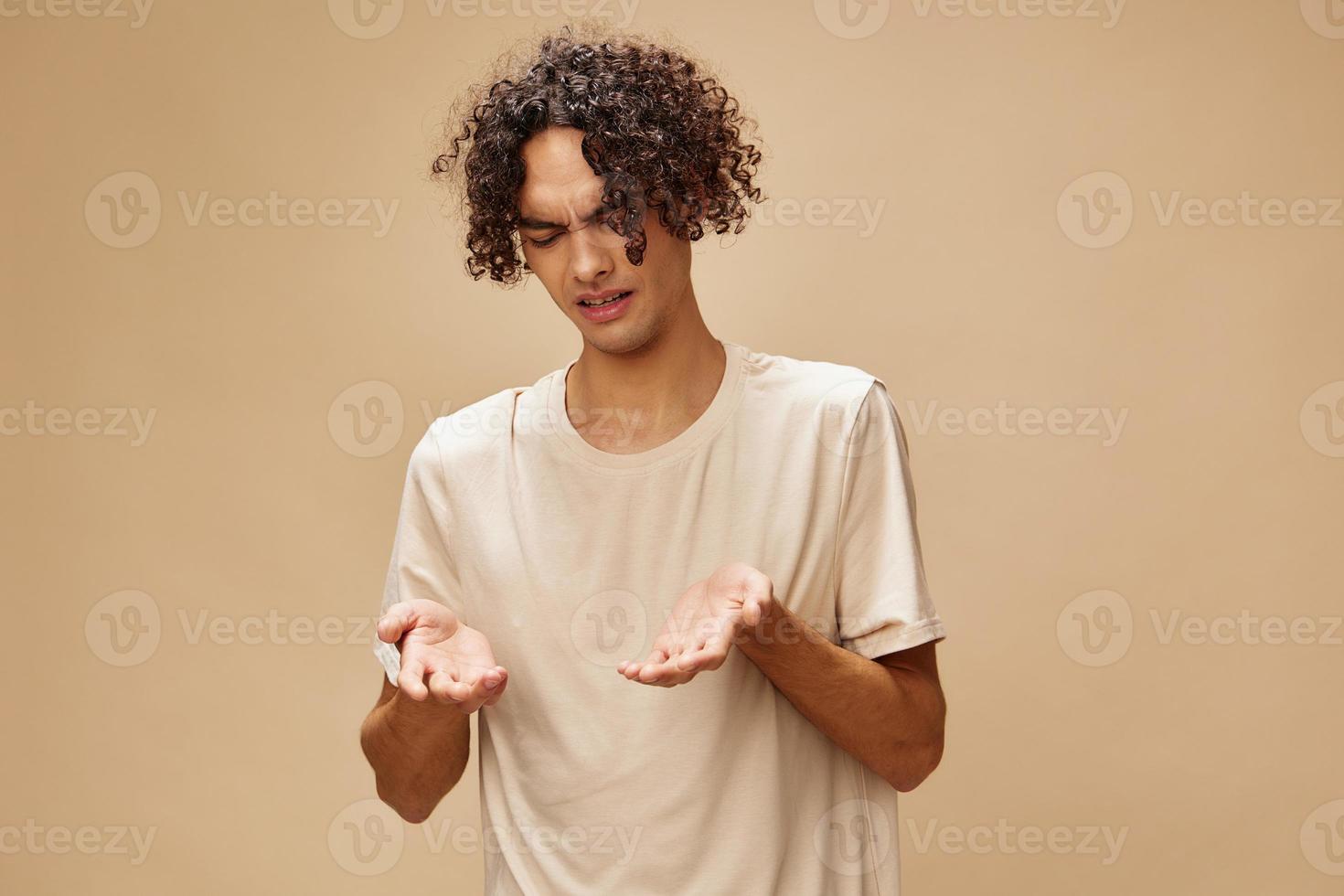 Unhappy discontented awesome tanned curly man in basic t-shirt with disgust looks aside posing isolated on beige background. Fashion New Collection offer. People Emotions concept. Free place for ad photo
