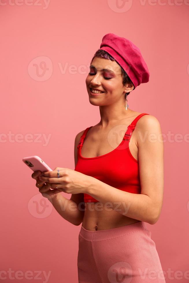 Young woman blogger with colored pink hair and short haircut flipping through the screen of the phone and typing a message with a smile in stylish clothes on a pink background photo