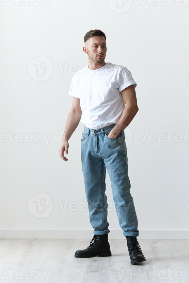 lleno cuerpo Disparo de hermoso grave bronceado hombre chico en básico camiseta sostiene mano en pantalones posando en blanco antecedentes. Moda estilo nuevo colección oferta. Copiar espacio para anuncio. modelado instantáneas foto