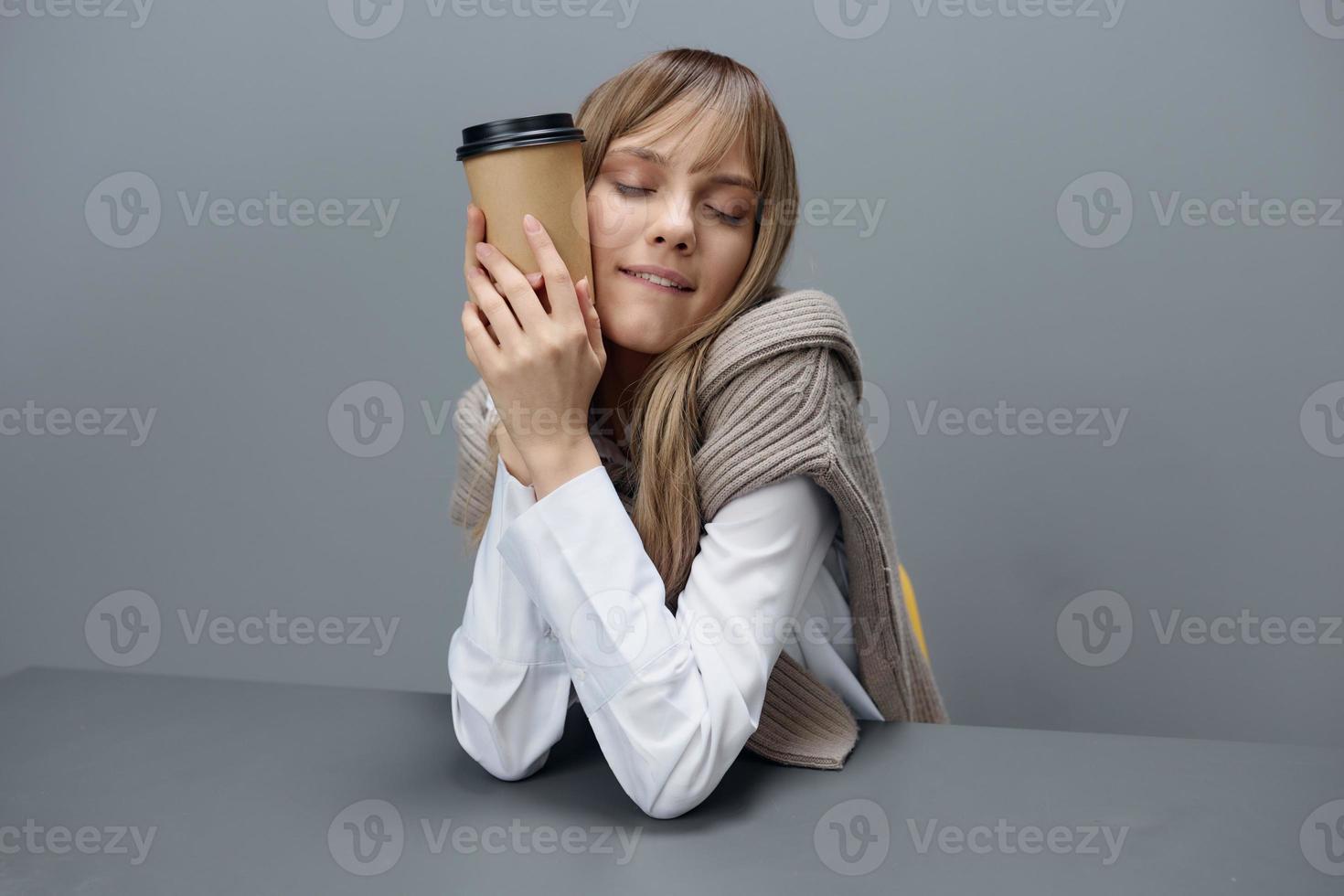 Cute happy young blonde student lady freelancer in warm sweater hug takeaway coffee sitting at workplace in gray modern home office. Coffee break Lover Concept. Copy space. Cool offer photo