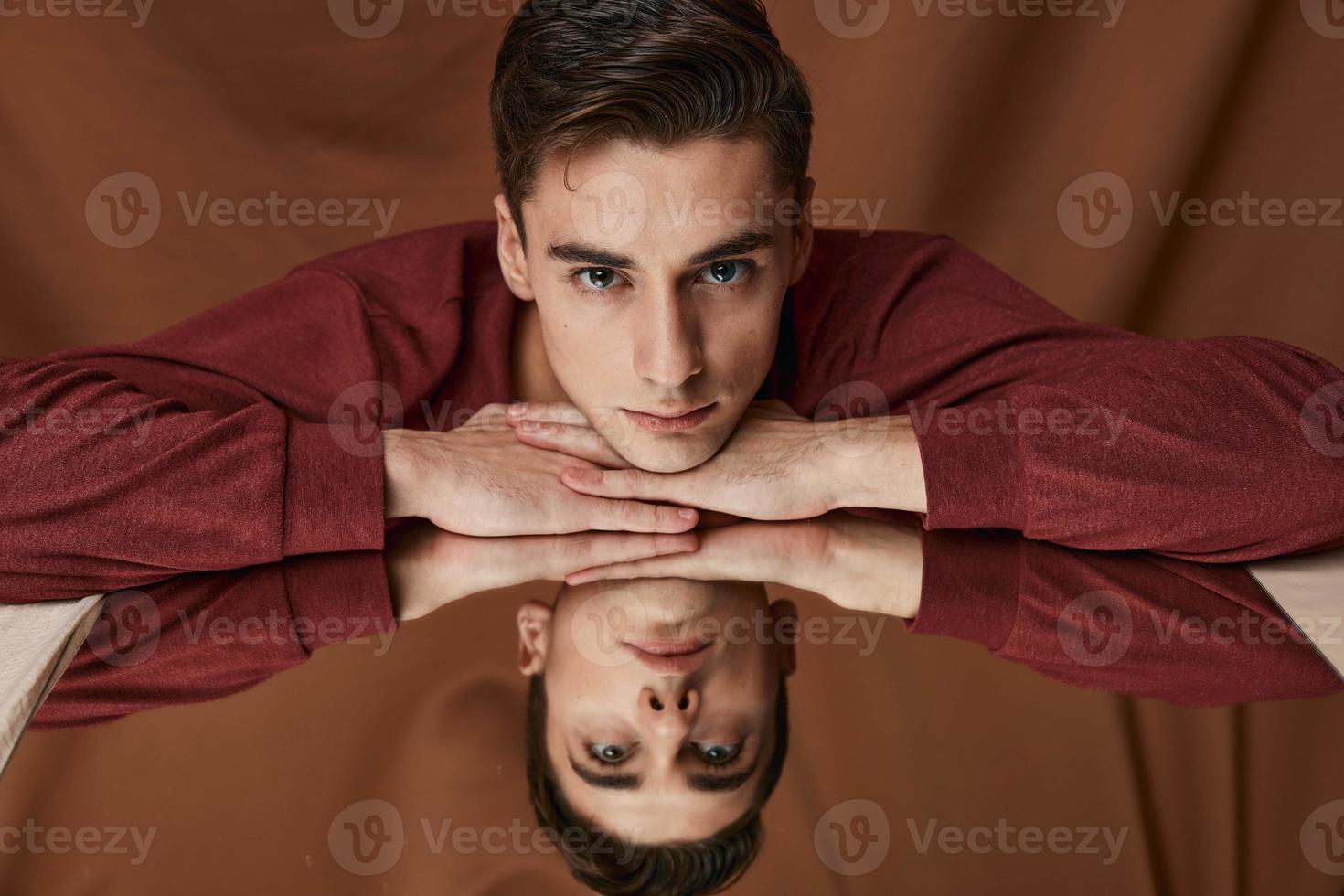 un hombre en un camisa es propensión en contra el vaso con un reflexión en el espejo en un marrón antecedentes foto