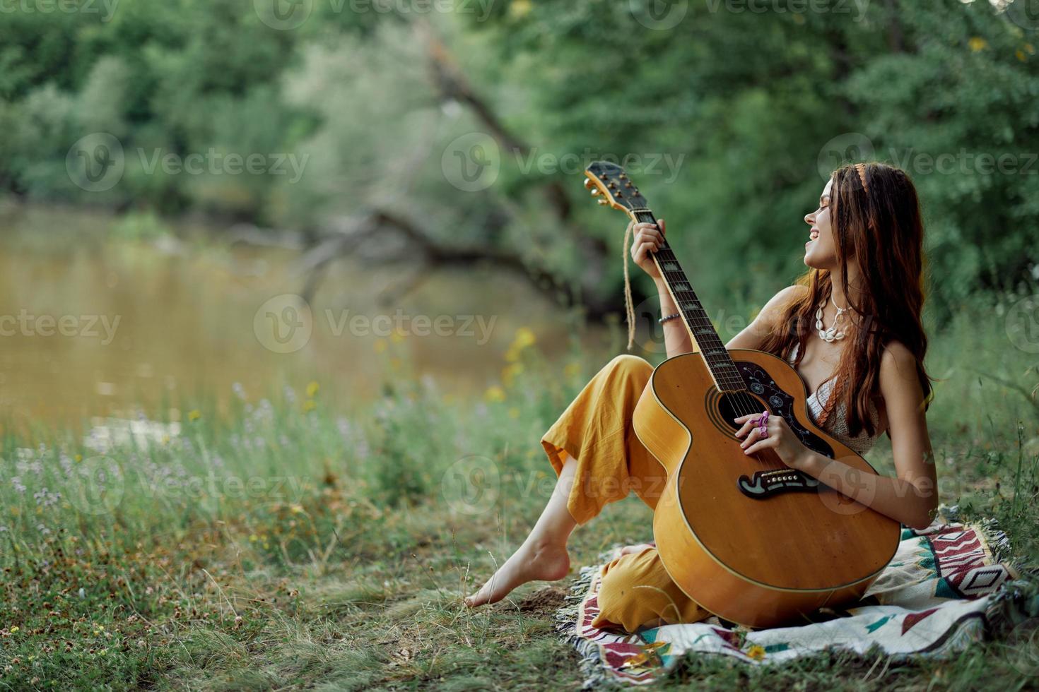 A hippie woman playing her guitar smiles and sings songs in nature sitting on a plaid in the evening in the sunset sunlight. A lifestyle in harmony with the body and nature photo