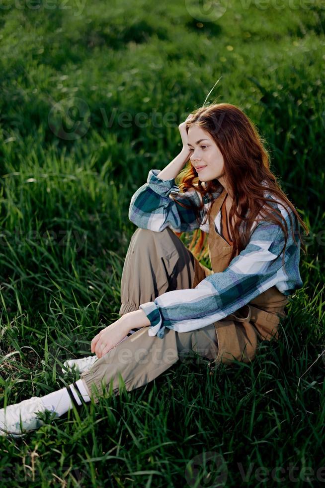 A young, beautiful woman lounges on the green grass in the park wearing sneaker pants and a plaid shirt and looks out into the setting sun photo