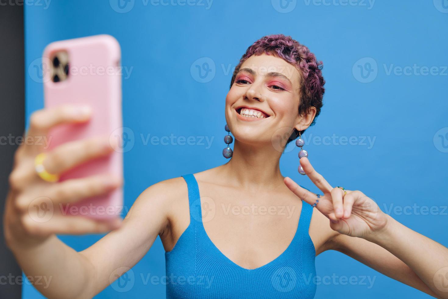 Young sports woman fashion blogger takes a picture of herself on the phone in blue sportswear smiling and showing her tongue on a blue monochrome background photo