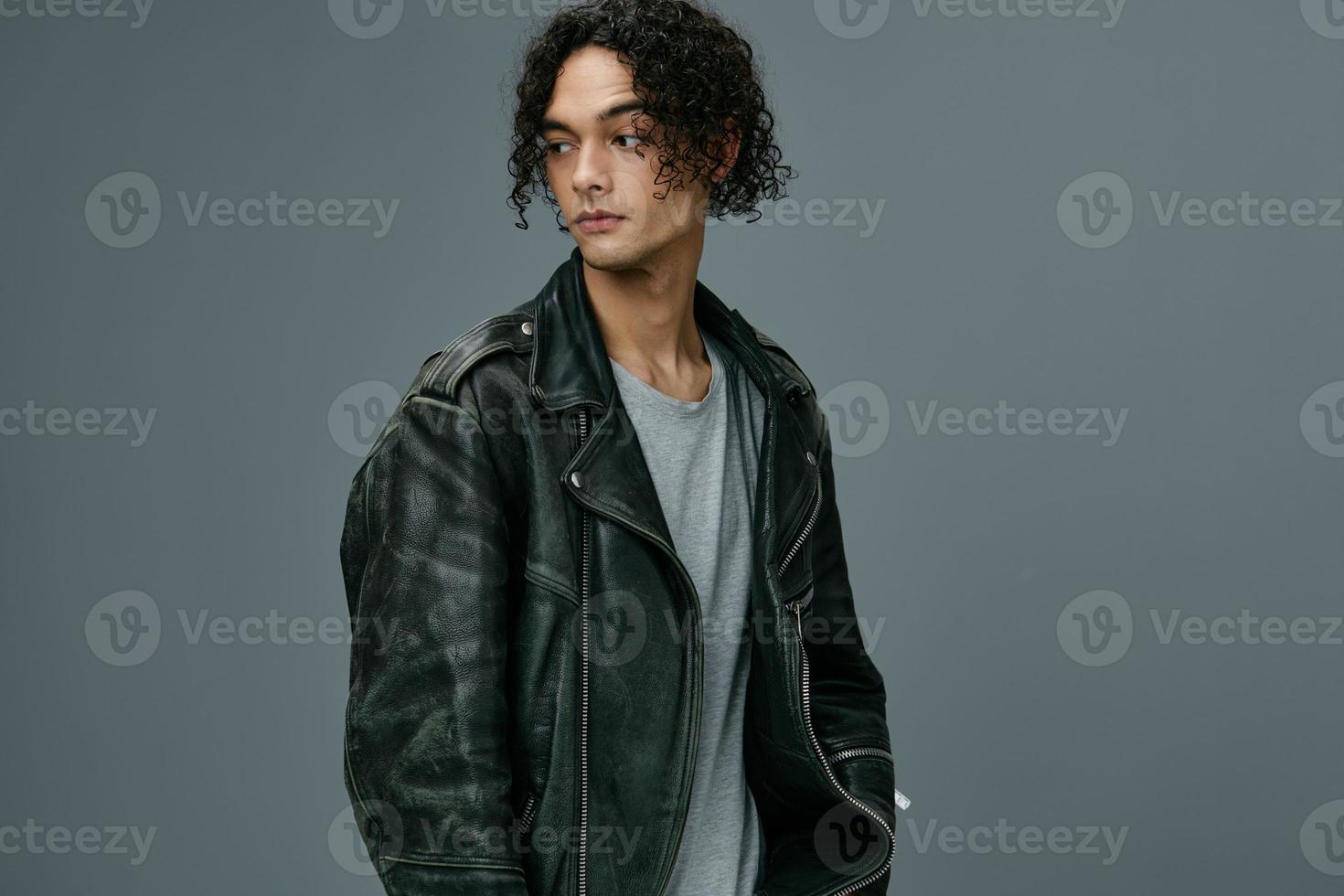 Handsome stylish tanned curly man leather jacket looks aside posing isolated on over gray studio background. Cool fashion offer. Huge Seasonal Sale New Collection concept. Copy space for ad photo