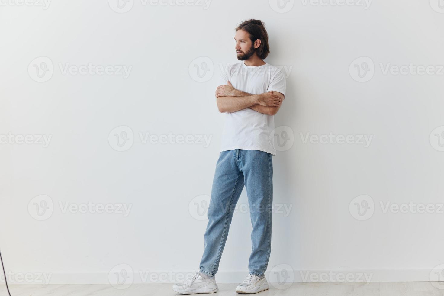 A man with a beard and long hair in a white T-shirt and blue jeans stands against a white wall leaning full-length on it, relaxed style photo