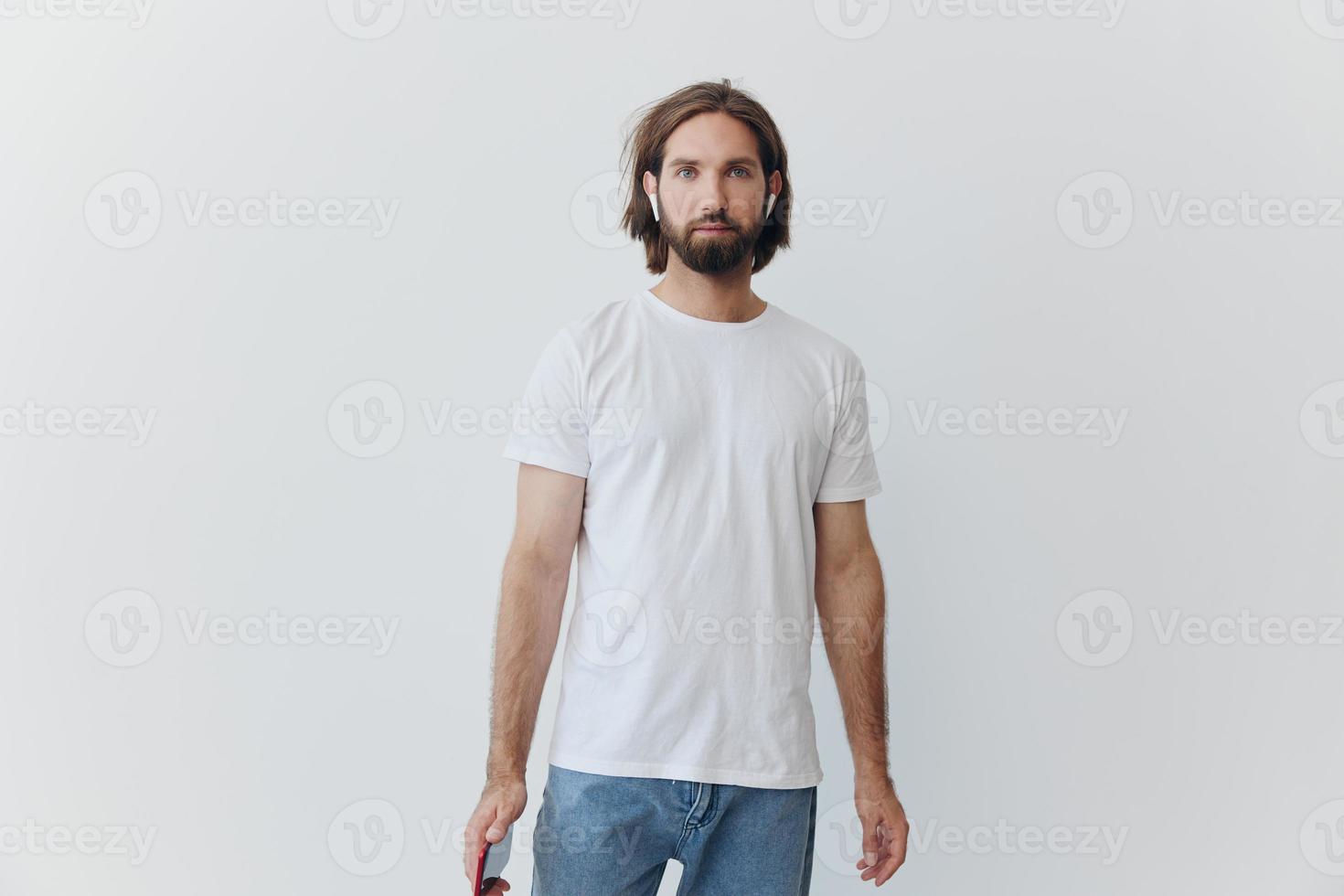 A man with a beard and long hair in a white T-shirt and blue jeans stands against a white wall, leaning against it and listening to music with wireless white headphones, staring thoughtfully photo