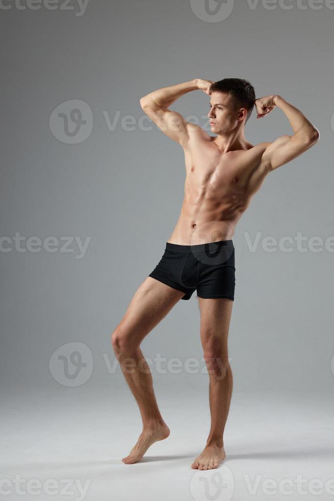 atleta en negro pantalones cortos sostiene manos detrás su cabeza desnudo torso gris habitación longitud total foto