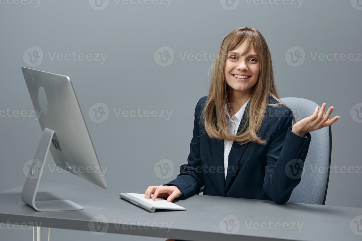 Smiling millennial blonde businesswoman worker in blue jacket using desktop computer look at camera in gray modern office. Remote Job, Technology And Career Profession Concept. Copy space photo