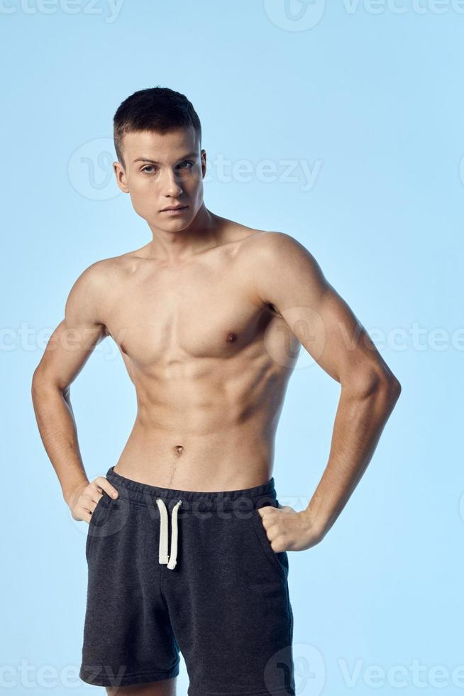 athlete in shorts holds his hands on the belt on a blue background inflated torso muscles of a bodybuilder photo