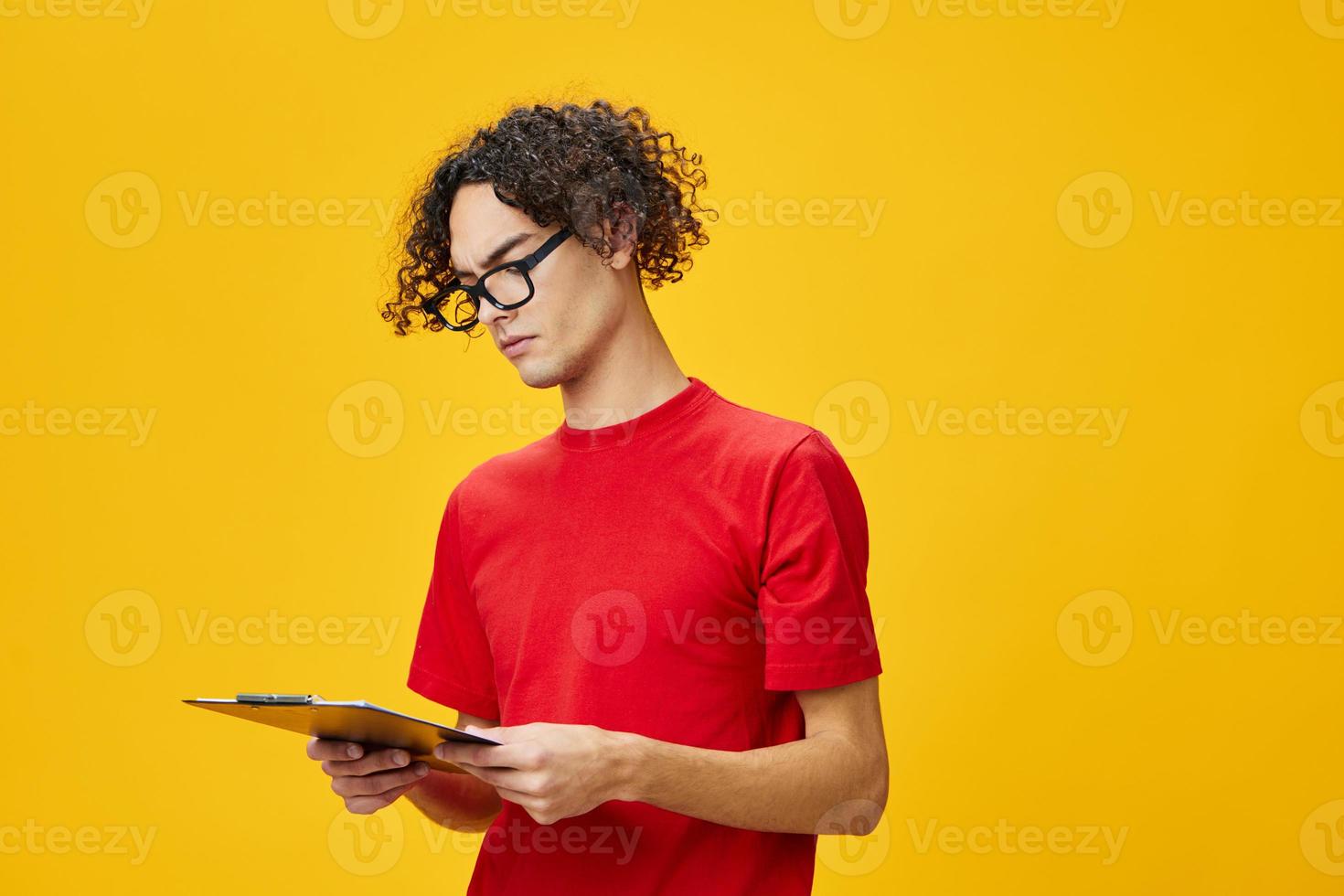 Thinking myopic young student man in red t-shirt funny eyewear holds tablet folder with study notes posing isolated on over yellow studio background. Free place for ad. Education College concept photo