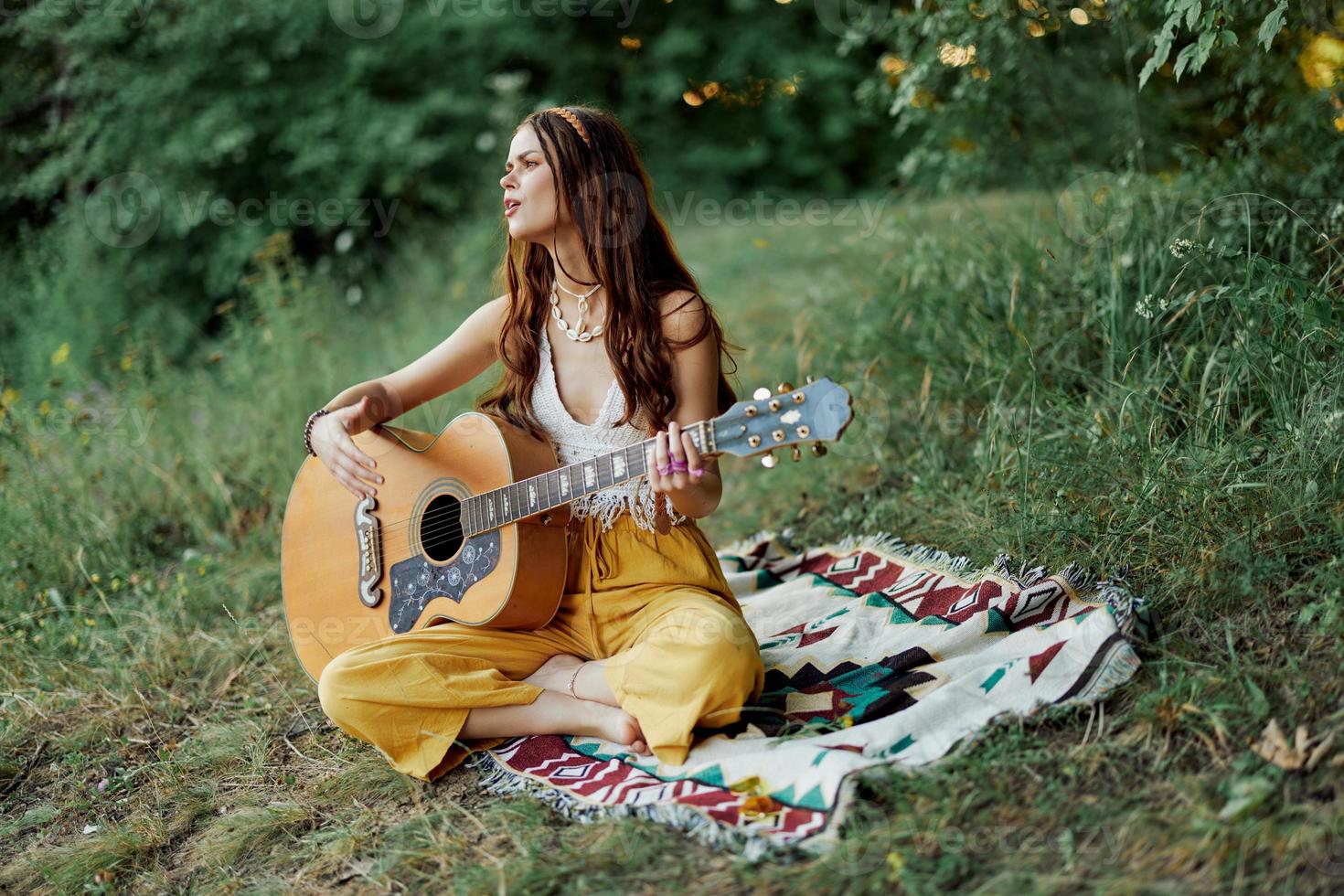 Young female hippie artist plays guitar and sings songs in eco-friendly clothing sitting on the ground outside in nature in the autumn looking out at the sunset photo