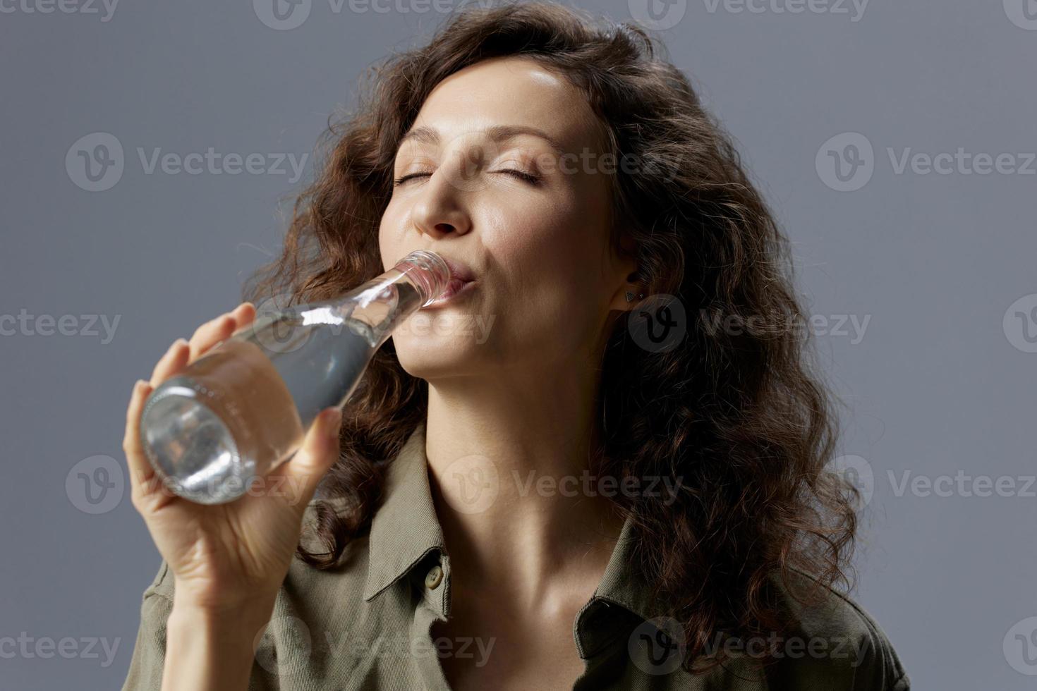 Enjoyed happy curly beautiful woman in casual khaki green shirt hold bottle drink clean mineral water posing isolated on over gray blue background. Healthy lifestyle. Water is Life concept. Copy space photo