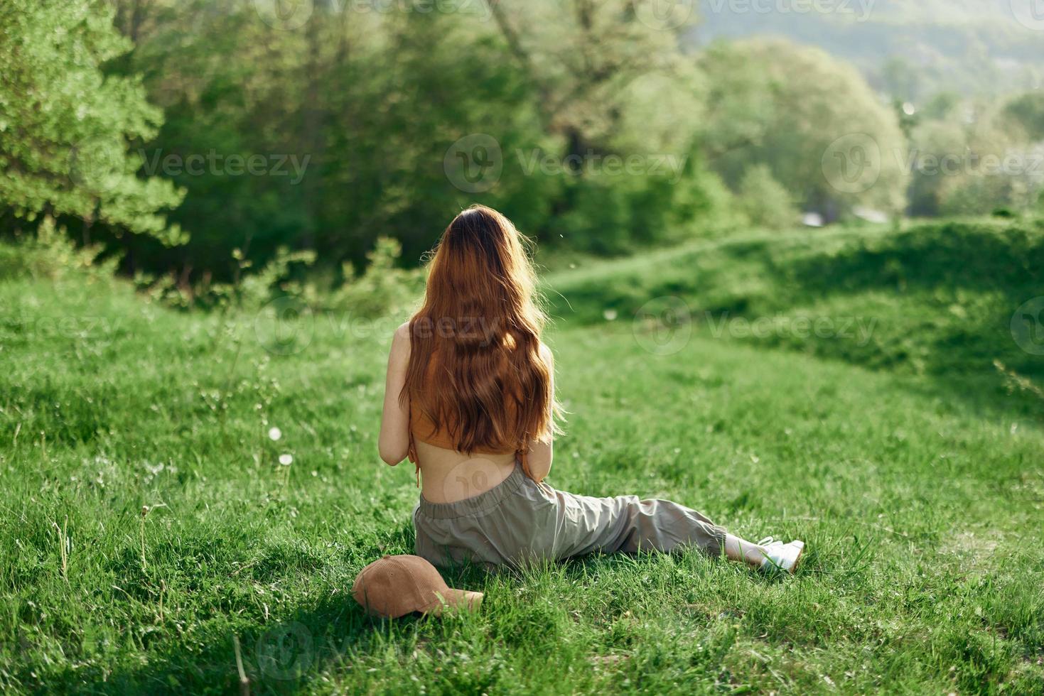 Top view of a woman in an orange top and green pants sitting on the summer green grass with her back to the camera with her phone, a young freelance student's concept of work and leisure photo