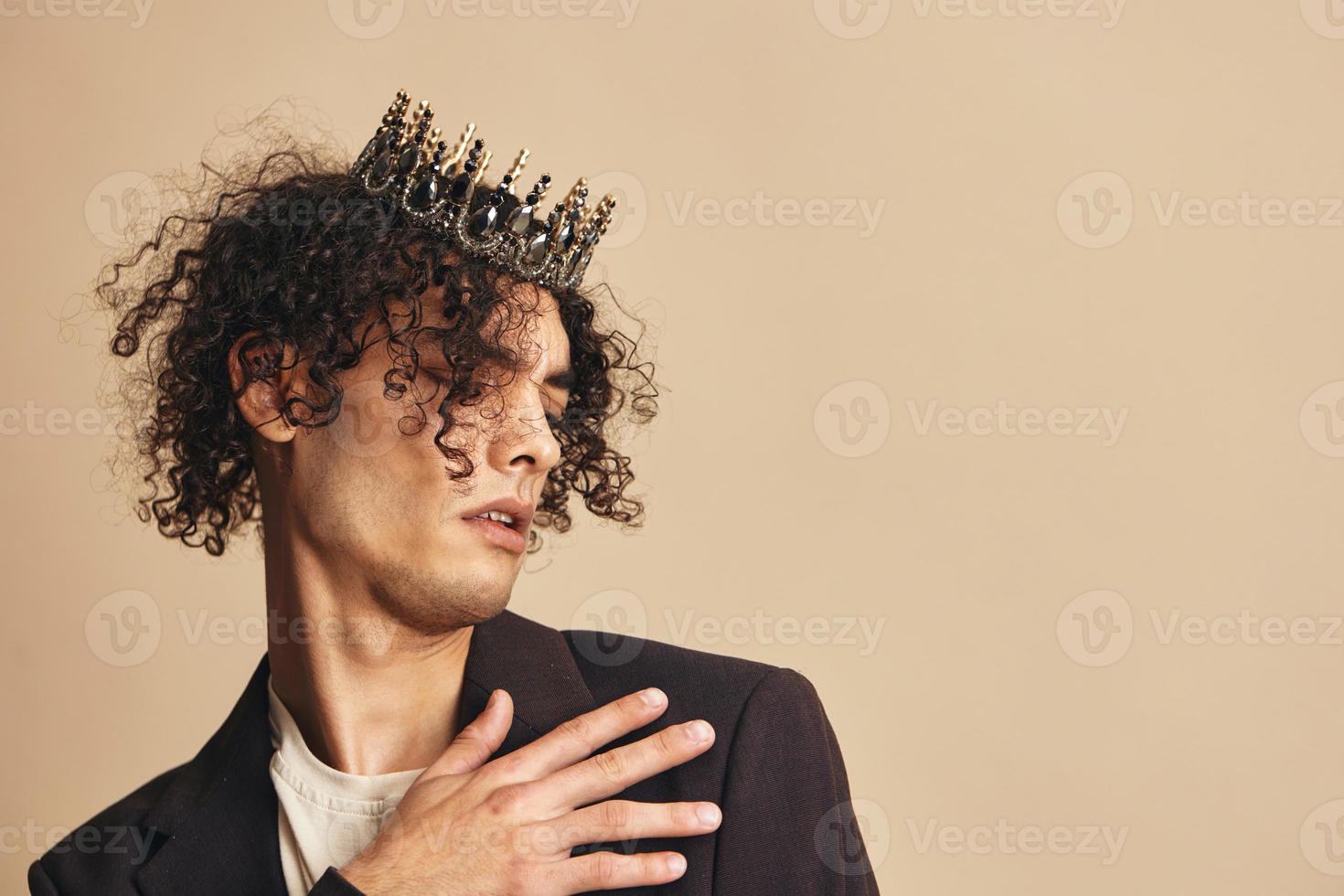 Stylish unhappy handsome funny narcissistic tanned curly man with crown posing isolated on over beige pastel background. Fashion New Collection offer. Retro style concept. Free place for ad photo