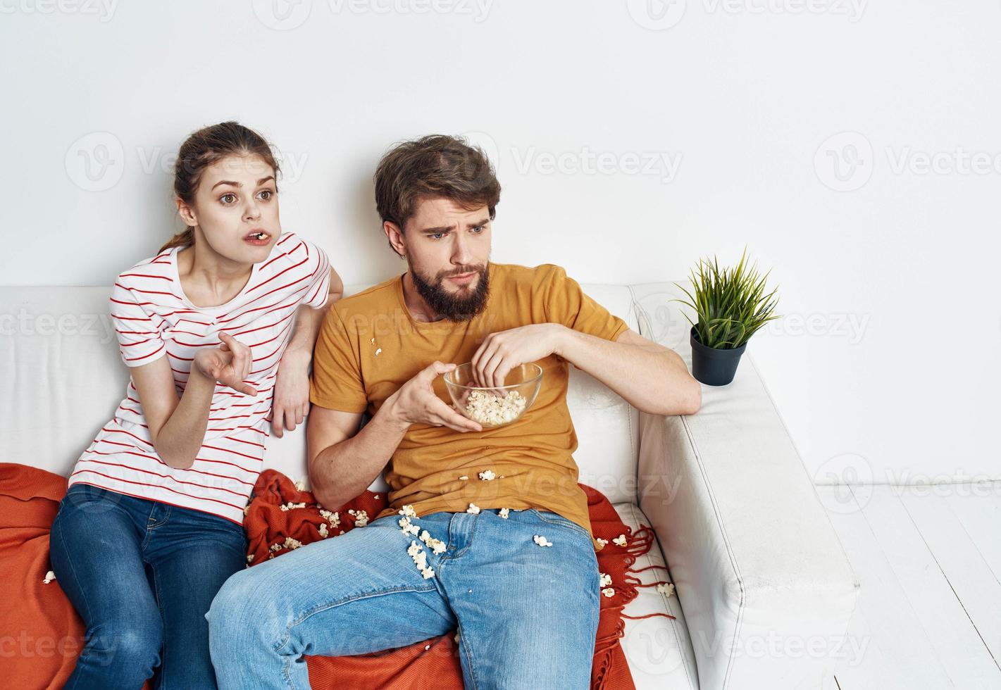 interesado mujer y barbado hombre sentar en el sofá palomitas de maiz en un plato y flor en un maceta foto