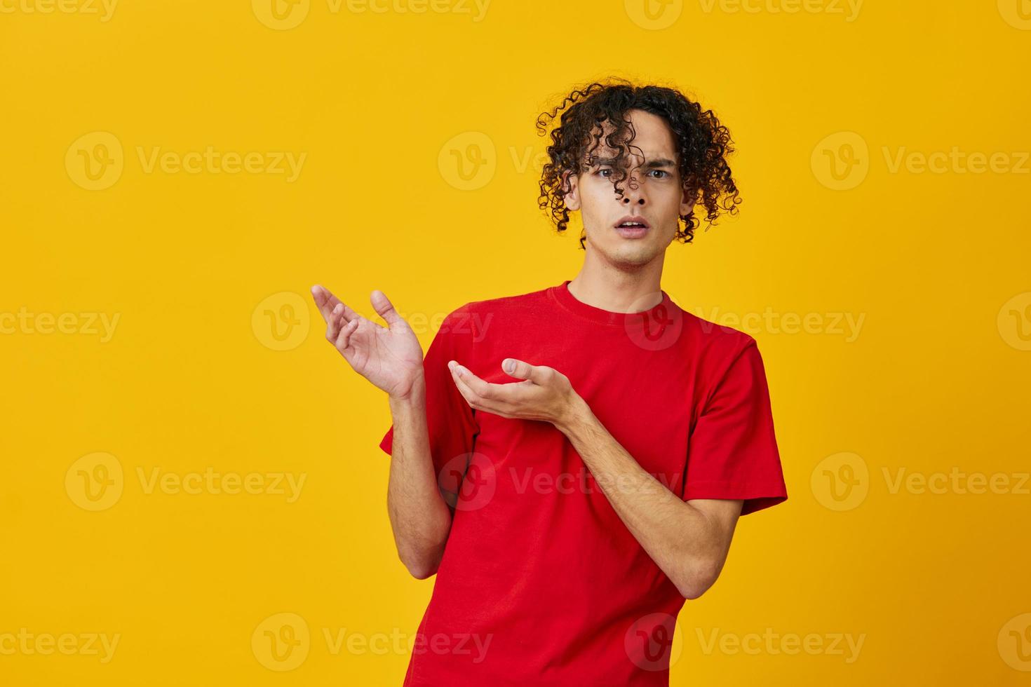 Confused funny Caucasian young man in red t-shirt point both hands aside posing isolated on over yellow studio background. The best offer with free place for advertising. Emotions for everyday concept photo