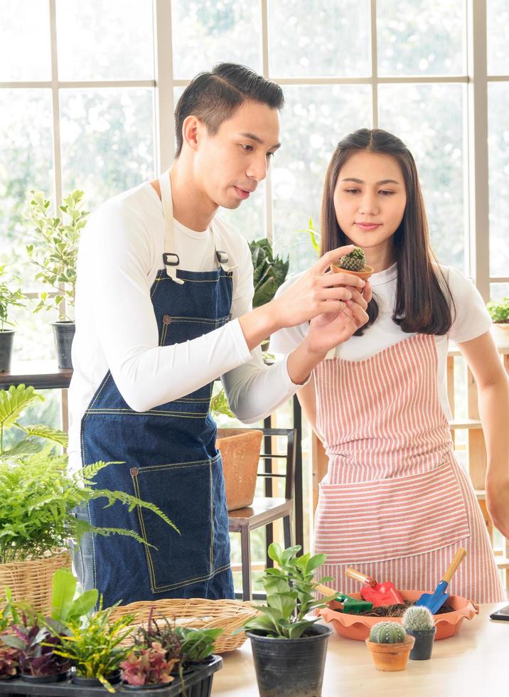 retrato amantes hermoso joven hombre bonito asiático mujer vistiendo blanco camiseta. y delantal bromas teniendo divertido ayuda organizar planta y agua plantas en pequeño ollas en el habitación arreglado plantas con amor felizmente foto