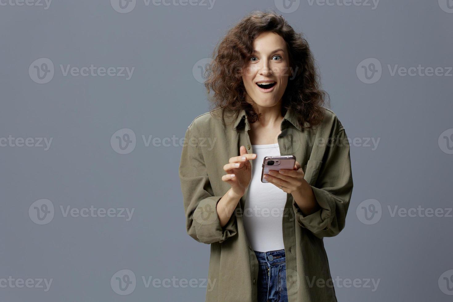 Shocked cheerful excited curly woman in casual khaki shirt received message about major sale of NFT project using phone posing isolated on gray background. Social Media Influencer concept. Copy space photo