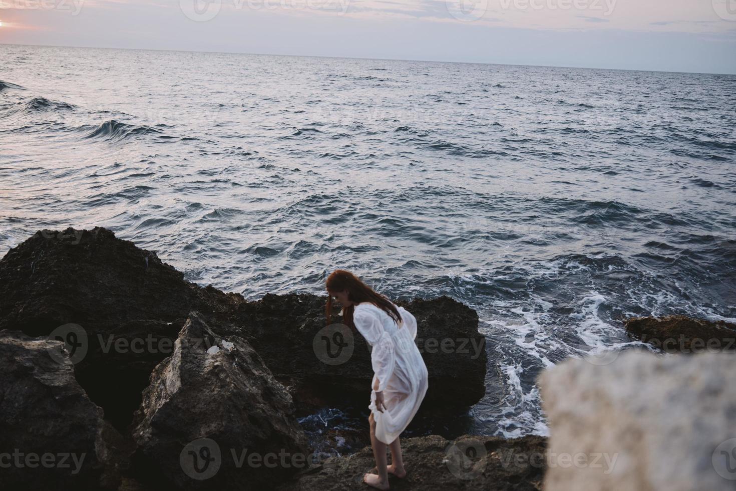 barefoot woman in a white dress with wet hair stands on a cliff photo