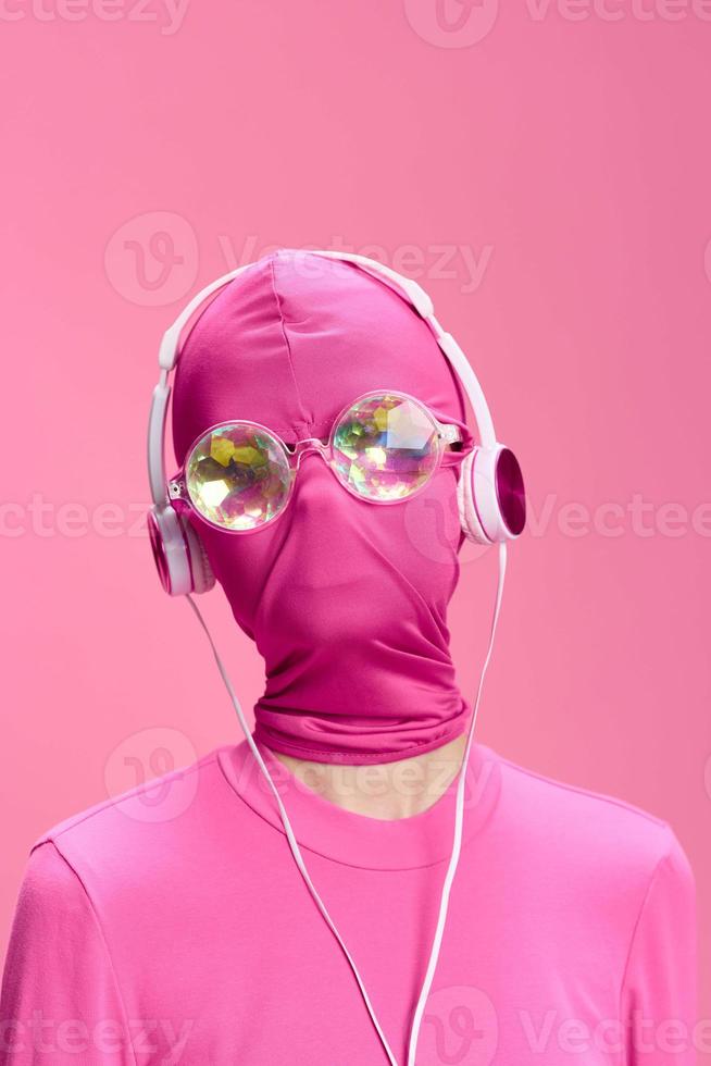 A crazy conceptual portrait of a young woman without a face mask on a pink background and wearing shiny sunglasses listening to music on her headphones. The concept of a new world and cyberspace photo