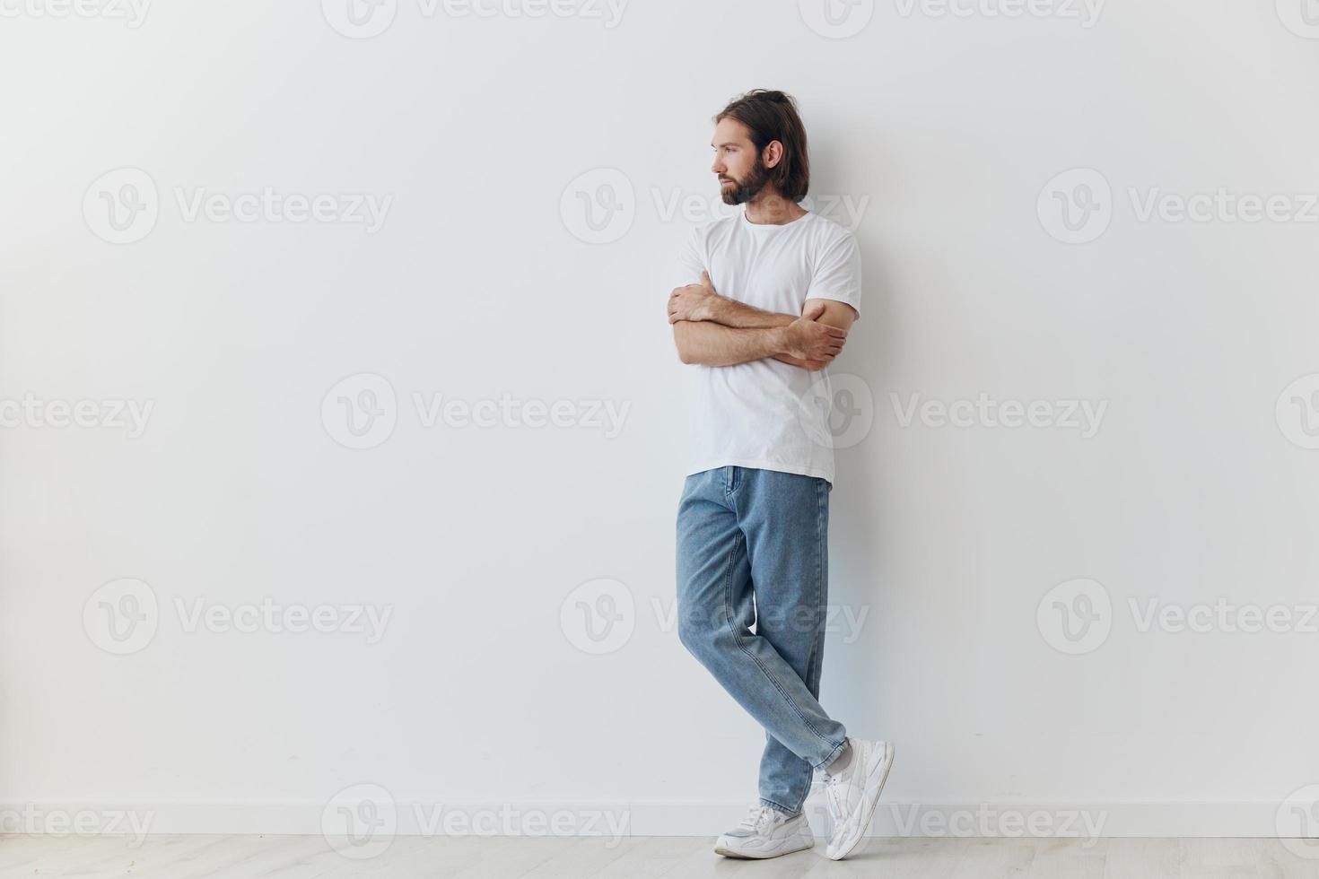 A man with a beard and long hair in a white T-shirt and blue jeans stands against a white wall leaning full-length on it, relaxed style photo