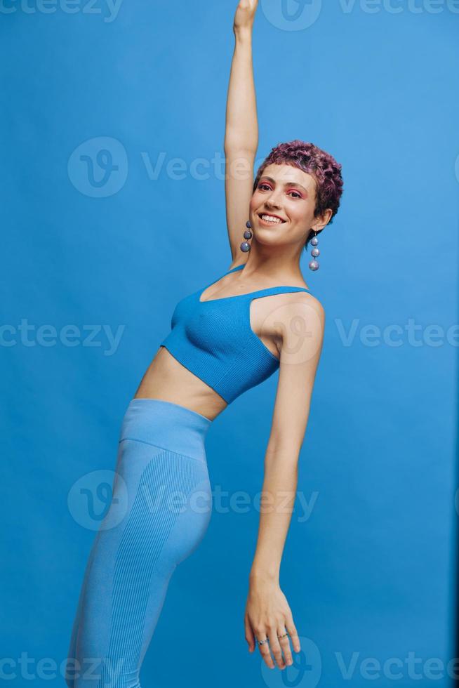 Young athletic fashion woman with colored hair and short haircut posing and dancing in blue sportswear smiling and looking at the camera on a blue monochrome background photo