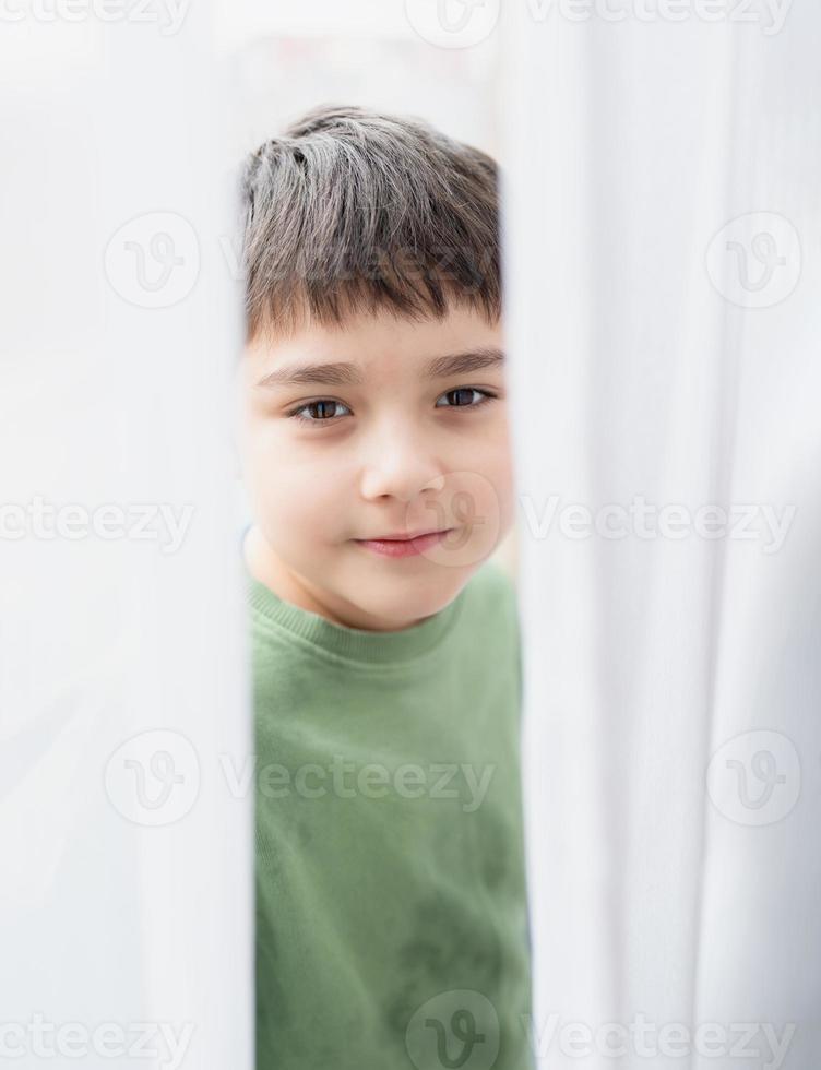 Happy kid with smiling face looks out from behind the curtains on window sill and plays at hide and seek ,Handsome young boy standing behind lace curtain with bright light in morning, photo