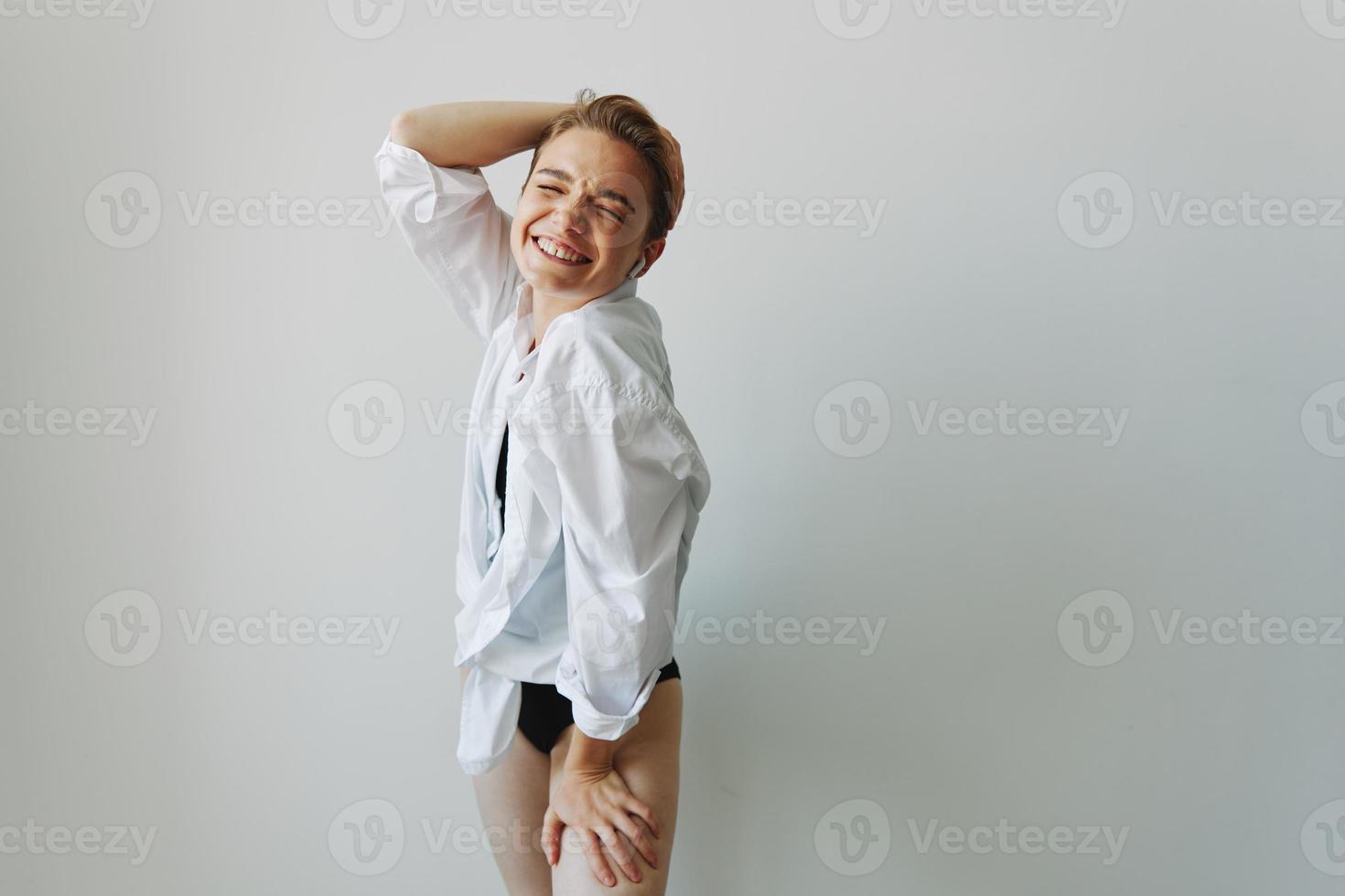 Young woman teenager listening to music with infertile headphones and dancing home, grinning with teeth with a short haircut in a white shirt on a white background. Girl natural poses with no filters photo