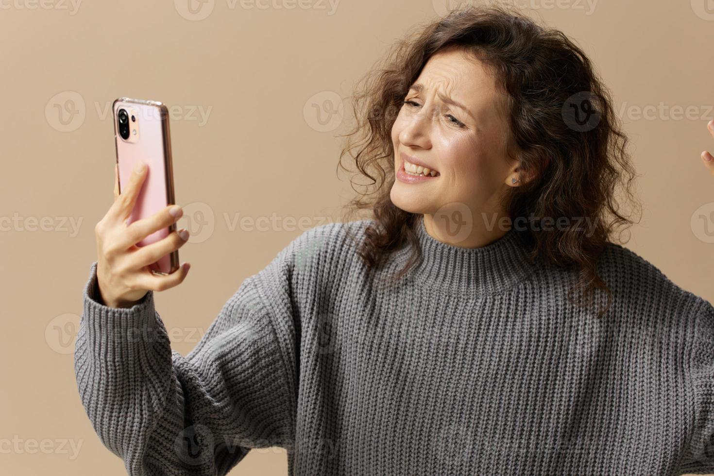 llorando escandalizando Rizado hermosa hembra en gris casual suéter hacer vídeo llamada con marido posando aislado en terminado beige antecedentes. social medios de comunicación, red, distancia comunicación concepto. Copiar espacio foto