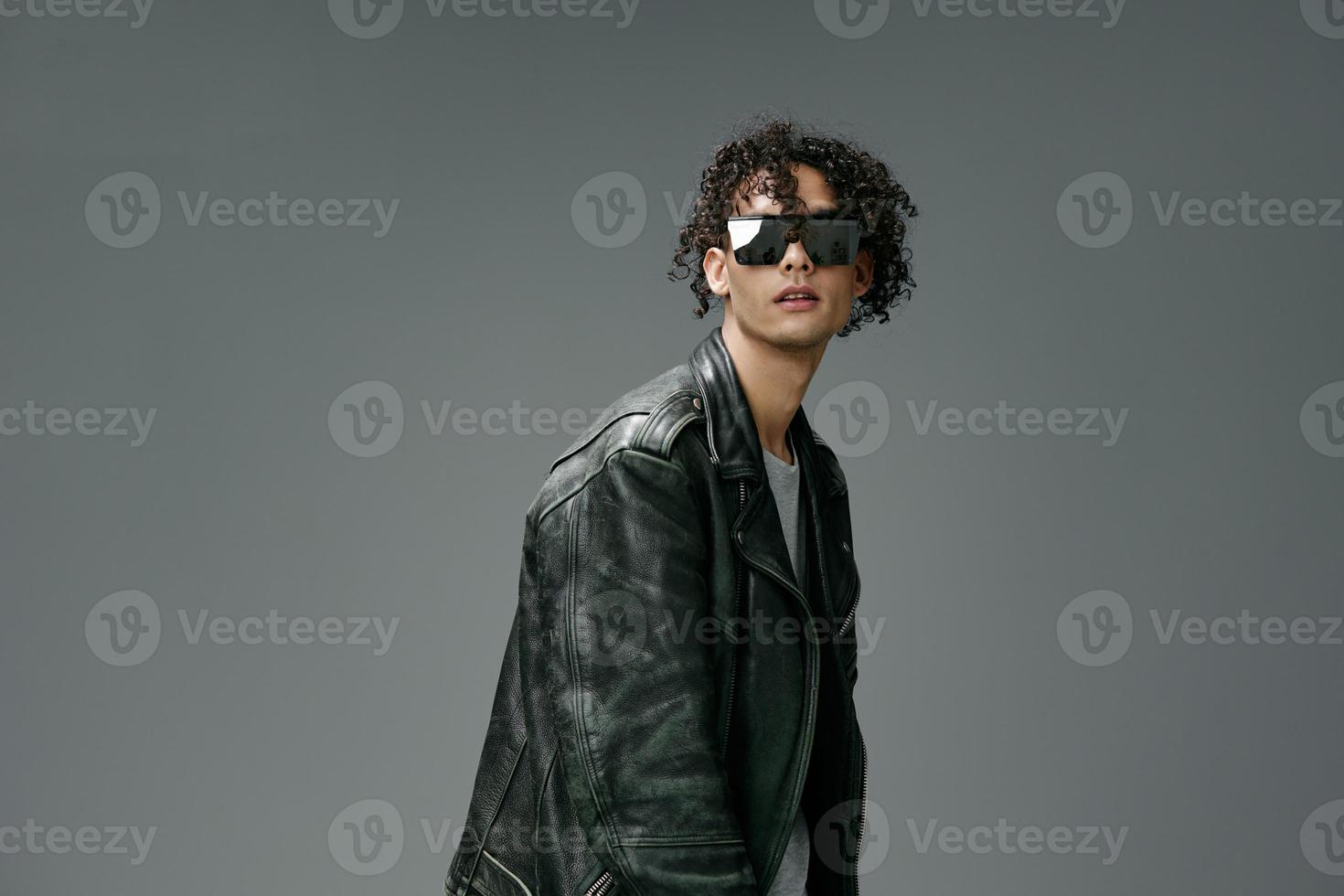 Stylish tanned curly man leather jacket trendy mirror sunglasses posing isolated on over gray studio background. Cool fashion offer. Huge Seasonal Sale New Collection concept. Copy space for ad photo
