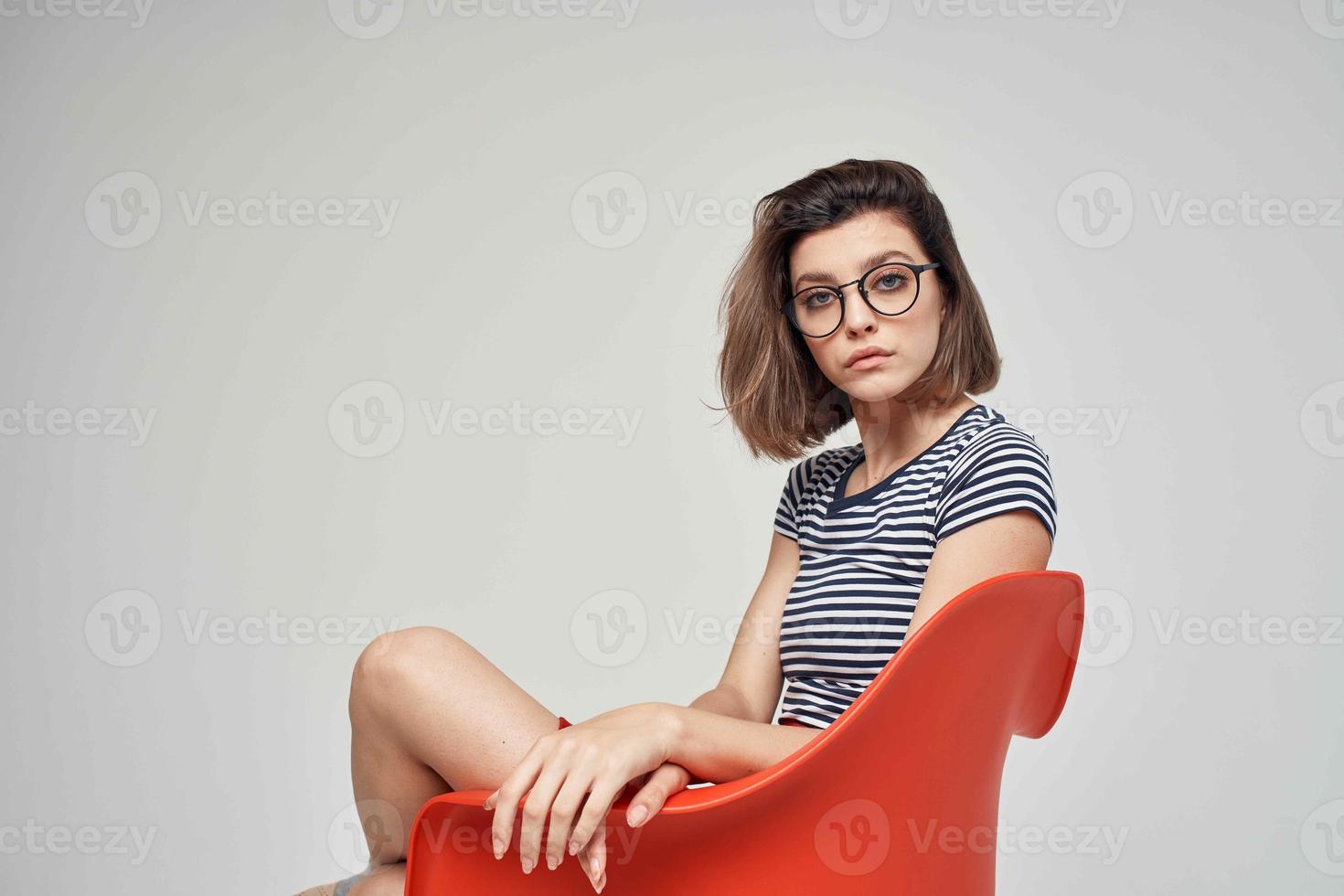 hermosa mujer en de moda ropa sentado en el rojo silla ligero antecedentes foto